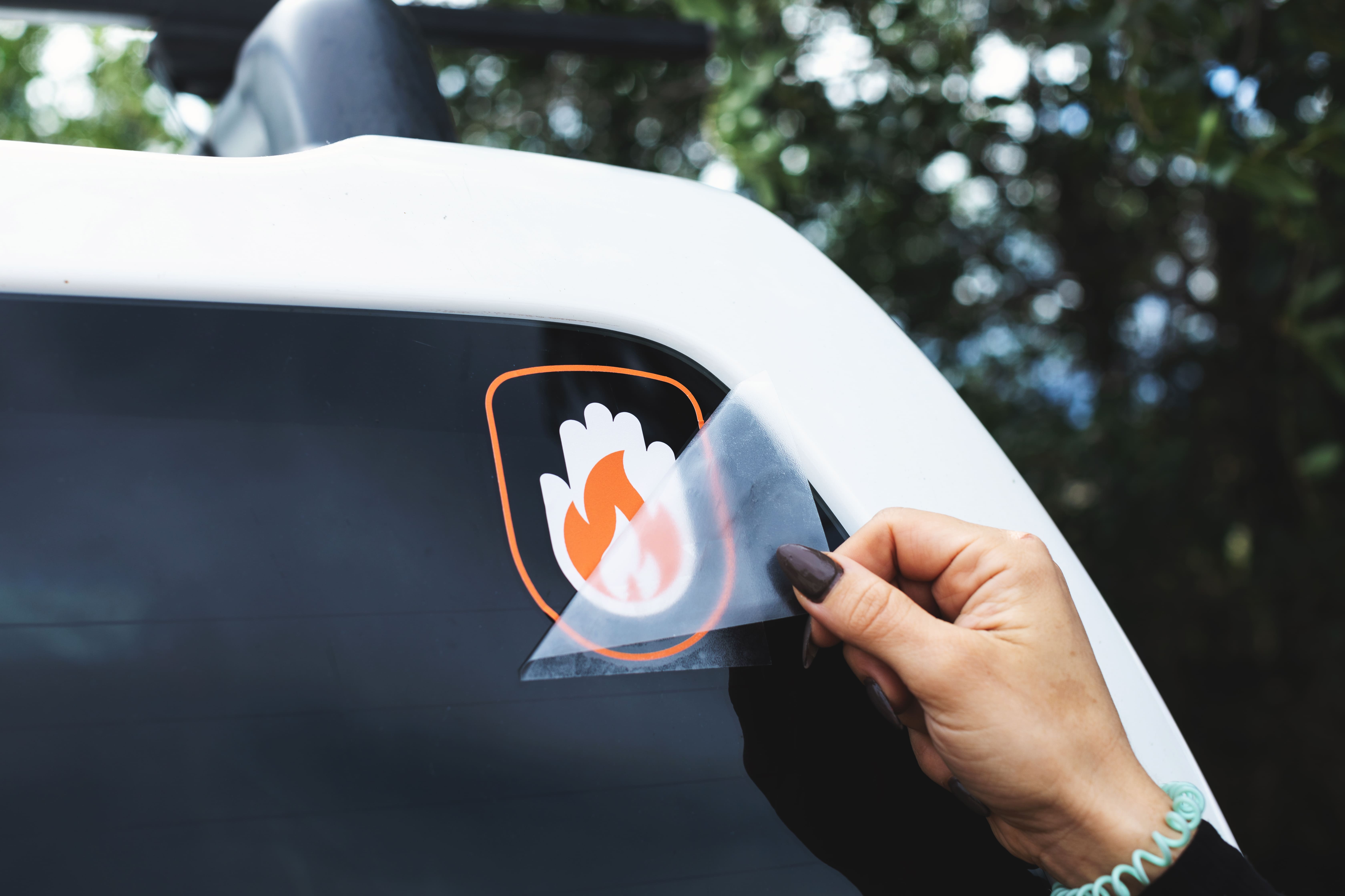 Printed transfer sticker in white and orange of a hand combined with a flame, applied to the rear window of a white car. A hand is peeling the transfer sheet off the sticker.