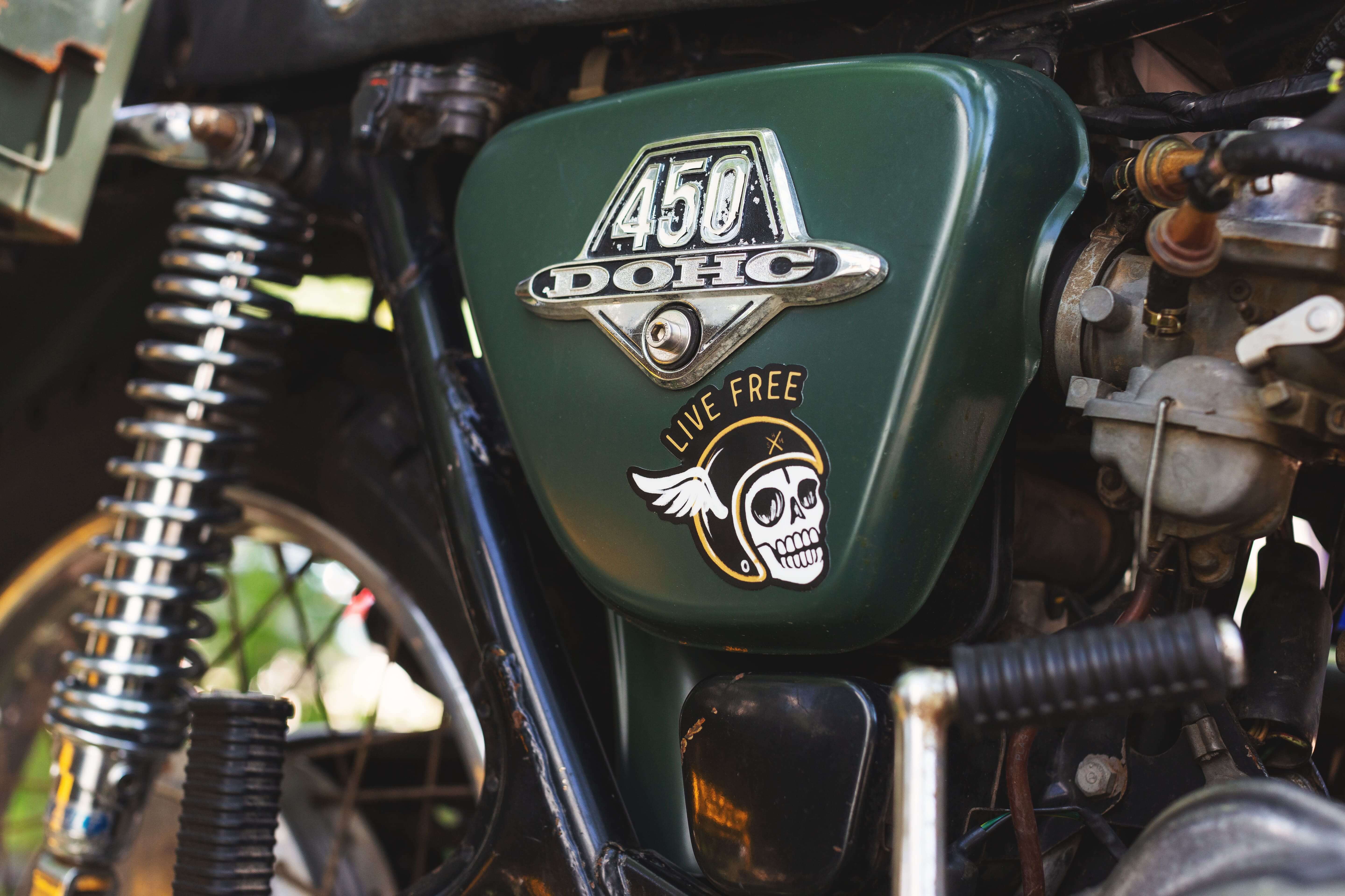 ‘Live Free’ die-cut sticker in a motorcycle style, showing a skull with a motor helmet, slapped on the tank of a green vintage motorcycle.
