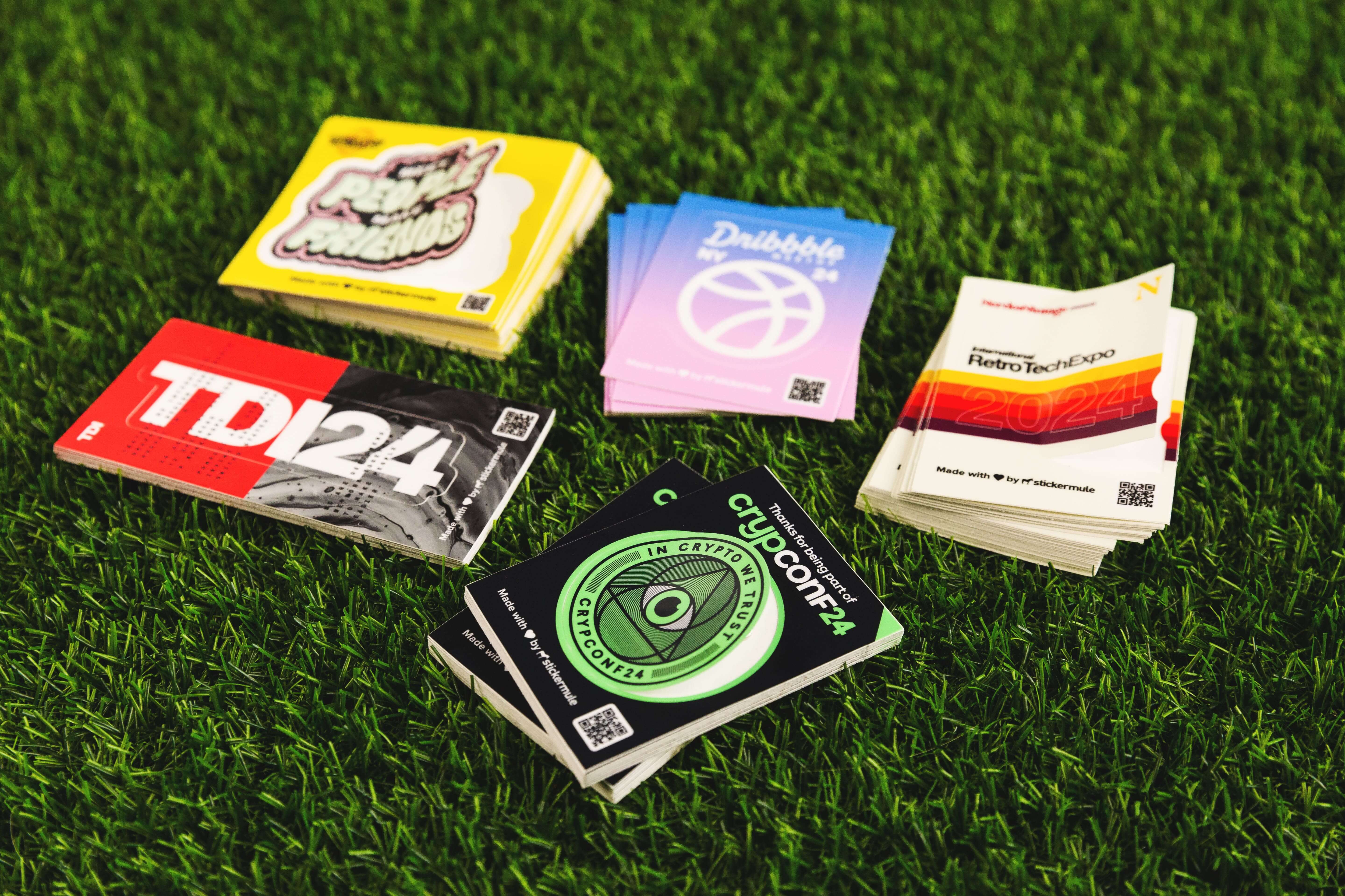 Stacks of unique event stickers on grass, featuring bold, custom designs for brands like CryptoConf, Dribbble, and RetroTechExpo.