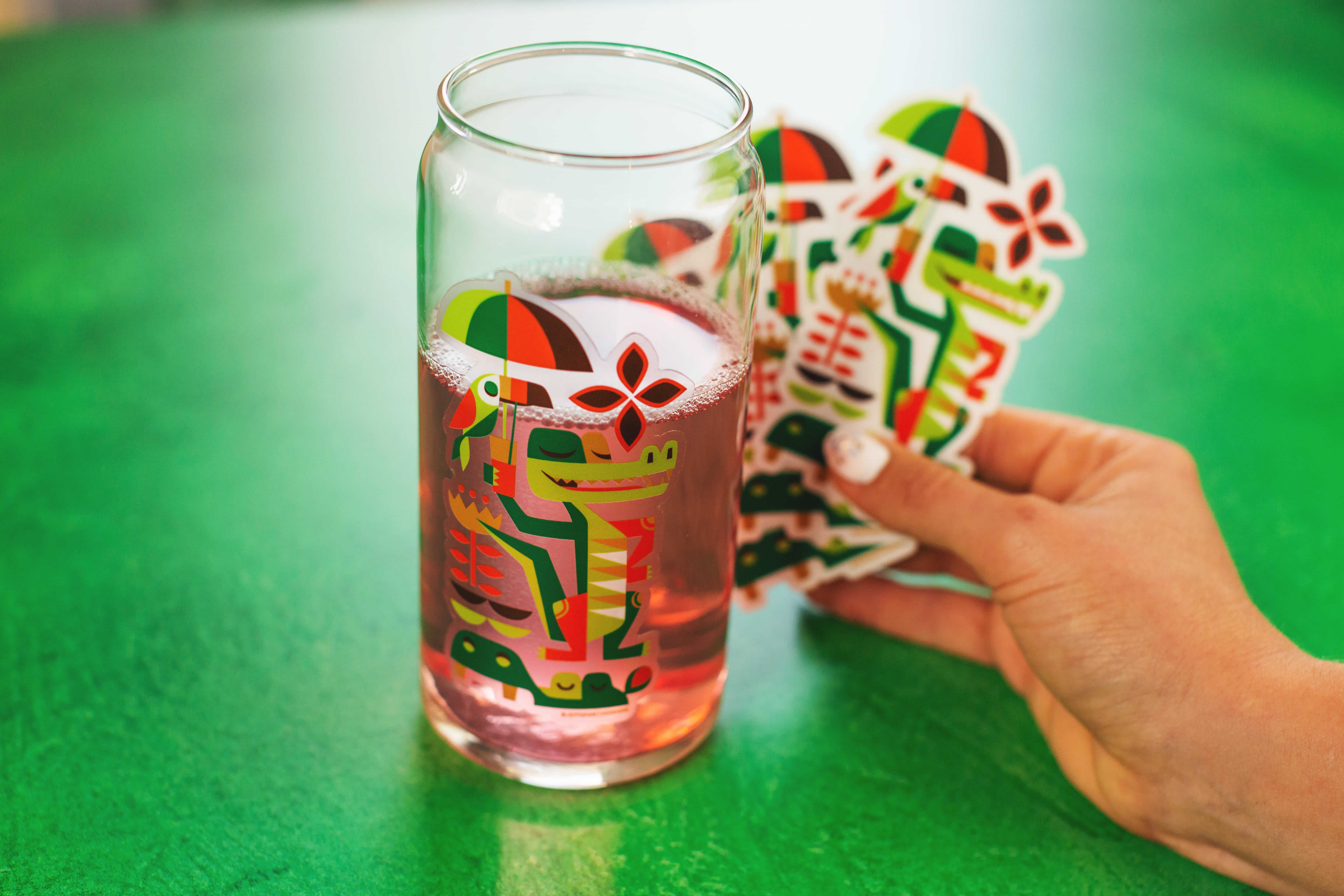 Um autocolante transparente de uma ilustração vetorial de um crocodilo a segurar um guarda-sol aplicado a um copo de limonada