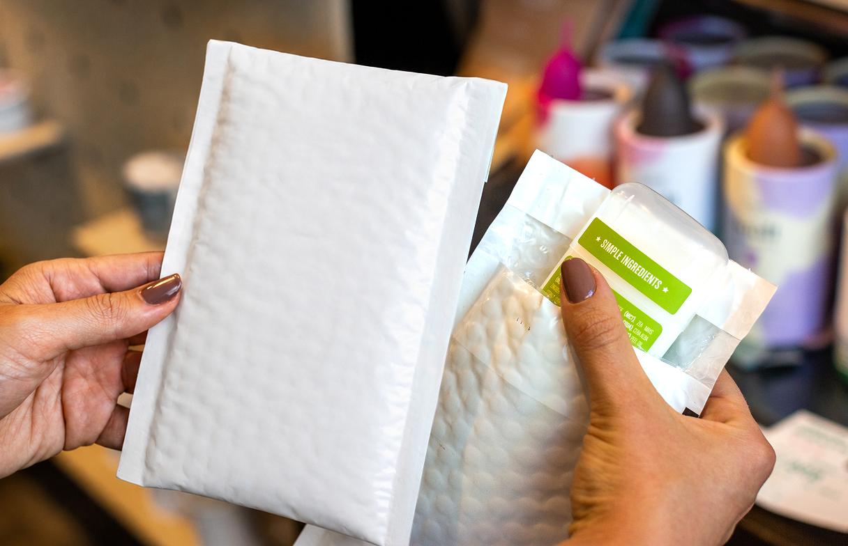 Hands holding a white padded bubble mailer next to a smaller clear bubble-wrapped package.