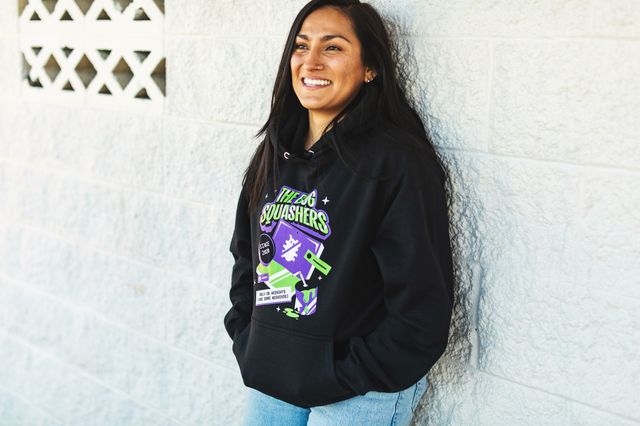 woman leaning against a wall in a black hoodie with a green and purple graphic on the front