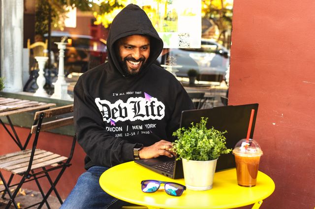 前面にグラフィックがプリントされた黒いパーカーを着た男性が、屋外のカフェでノートパソコンをタイピングしている